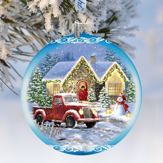 A Christmas ornament depicting a red pickup truck parked in front of a snow-covered house, with a snowman and Christmas lights visible.