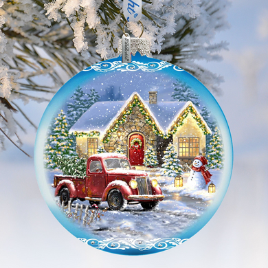 A Christmas ornament depicting a red pickup truck parked in front of a snow-covered house, with a snowman and Christmas lights visible.