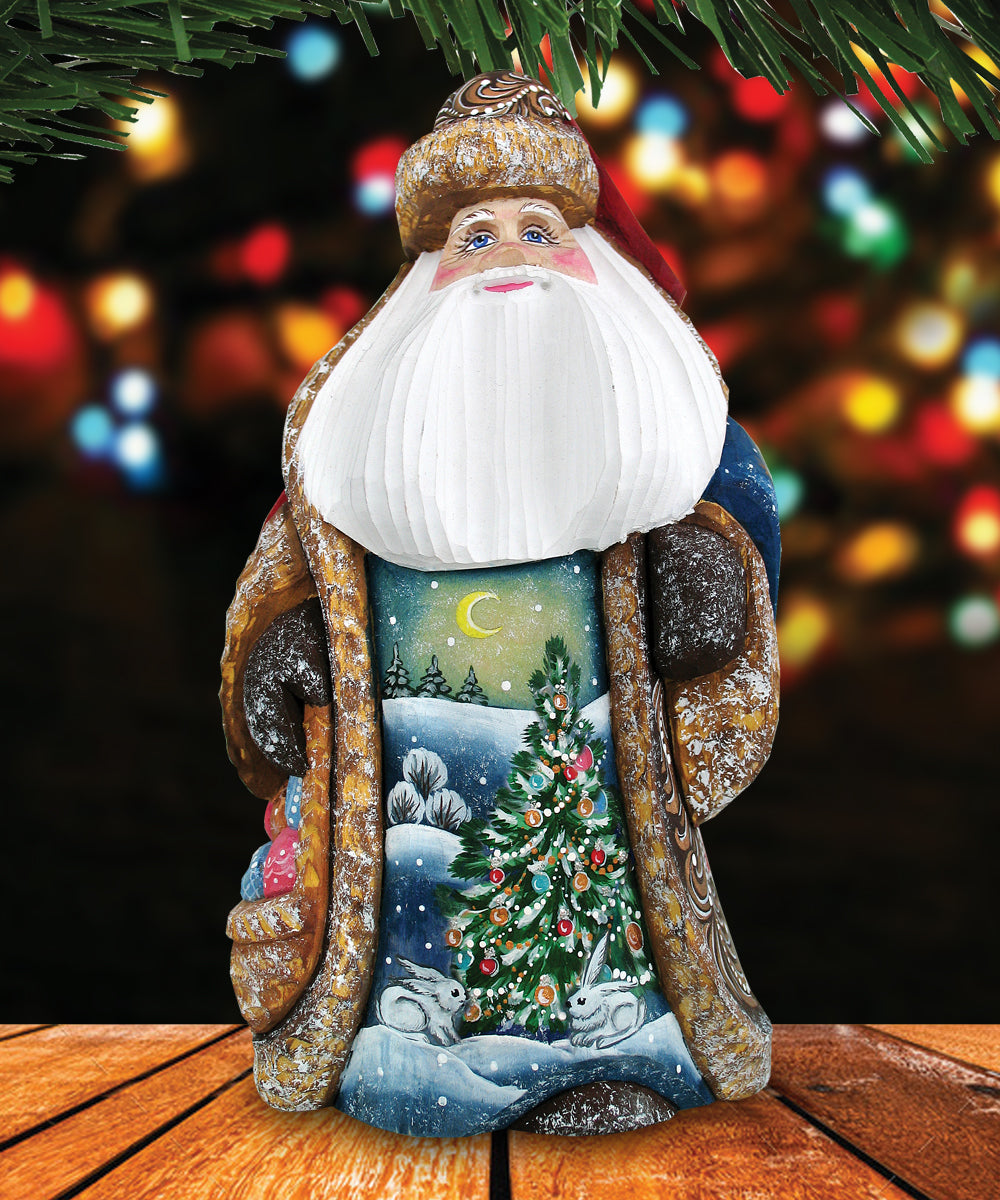 A figurine of Santa Claus with a white beard and hat, wearing a blue and gold outfit, is sitting on a wooden surface with a snowy landscape and Christmas tree in the background.