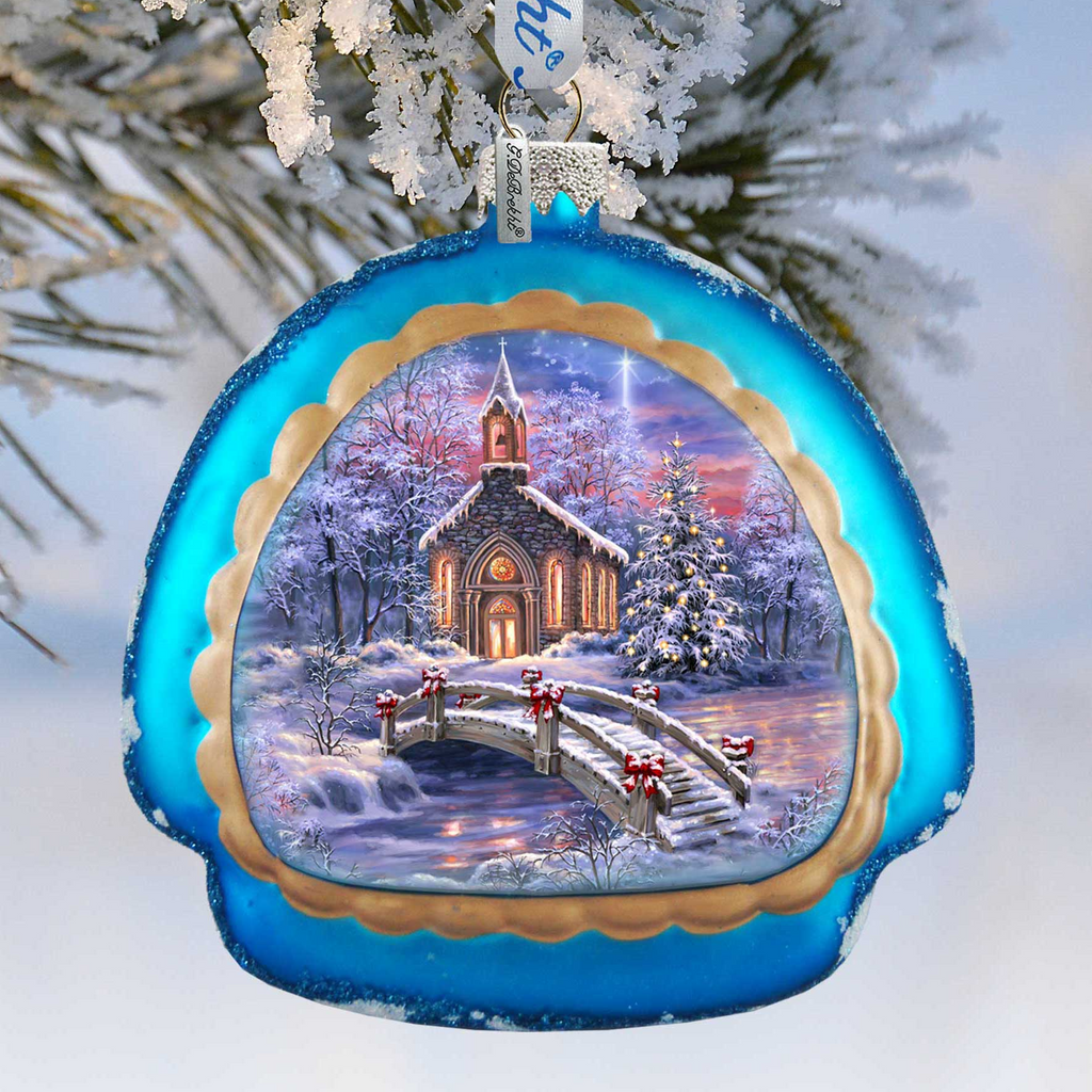 A blue ornament with a winter scene depicting a church, a bridge, and snow-covered trees.