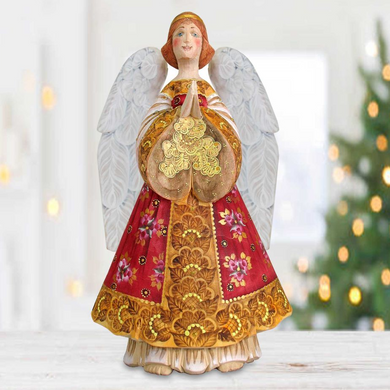 A figurine of an angel with wings and a red and gold dress, holding a flower in its hands, stands in front of a blurred Christmas tree.