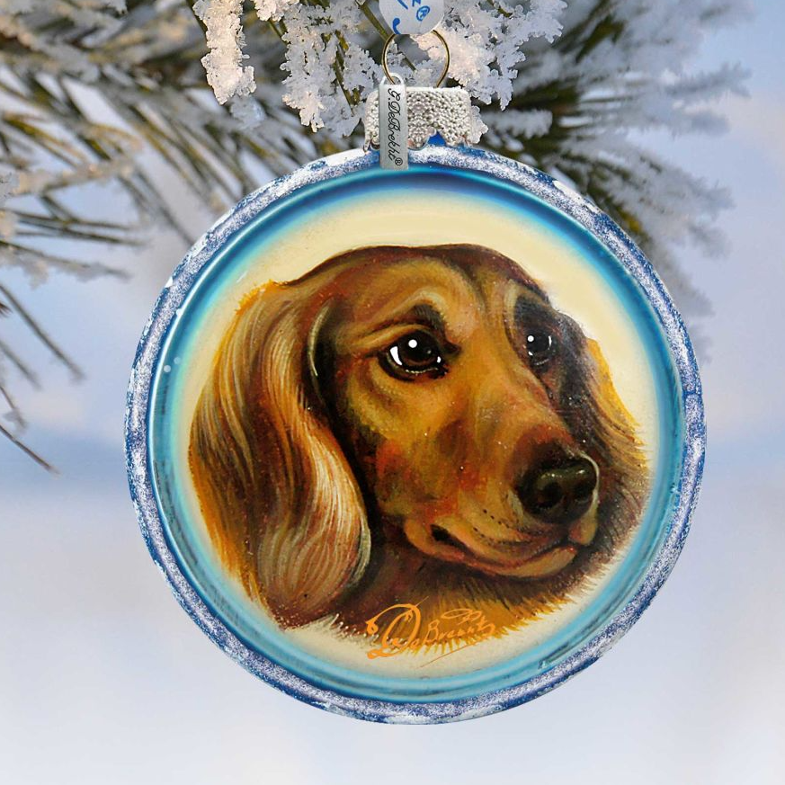 A blue ornament with a painting of a dog's face hanging from a branch of a snow-covered tree.