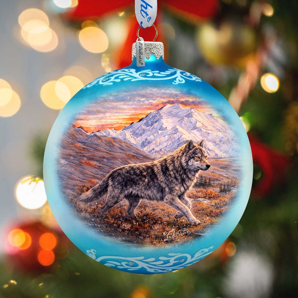 A blue Christmas ornament with a painting of a wolf in a snowy mountain landscape.