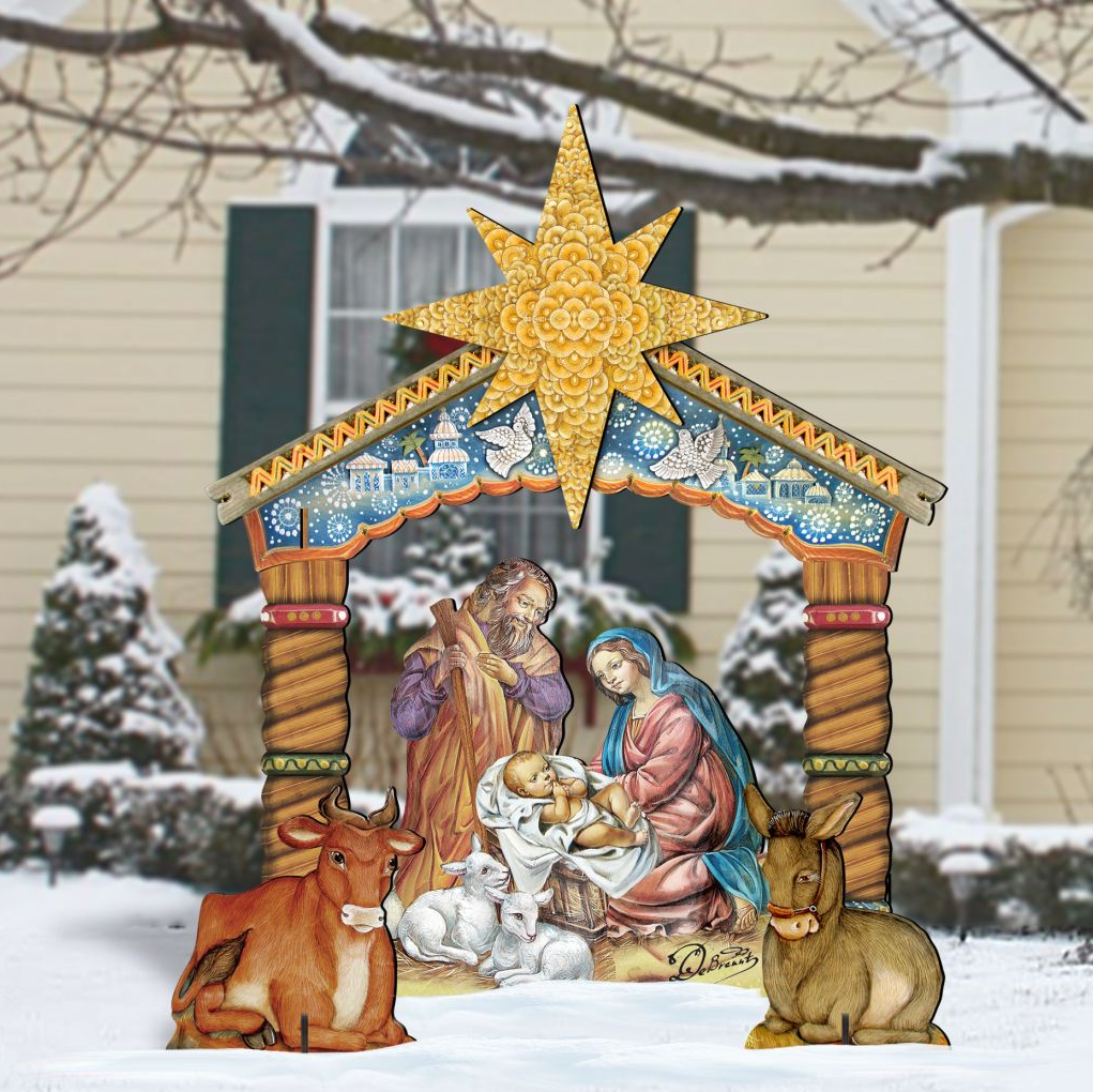 A large, colorful, and detailed cardboard nativity scene depicting the birth of Jesus Christ, with figures of the Virgin Mary, baby Jesus, and other figures arranged in a manger. The scene is set against a snowy background, with a house and trees visible in the distance.