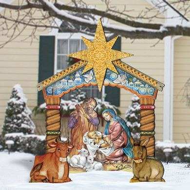 A large, colorful, and detailed cardboard nativity scene depicting the birth of Jesus Christ, with figures of the Virgin Mary, baby Jesus, and other figures arranged in a manger. The scene is set against a snowy background, with a house and trees visible in the distance.