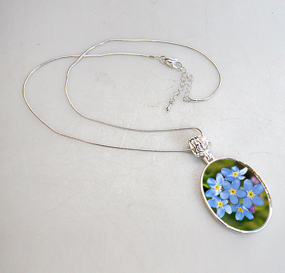A silver necklace with a pendant featuring a close-up image of blue forget-me-not flowers.