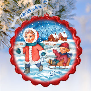 A red ornament with a snowy winter scene featuring two children playing in the snow, one on a sled and the other walking.