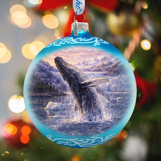 A blue ornament with a painting of a whale leaping out of the water, set against a snowy mountain landscape.