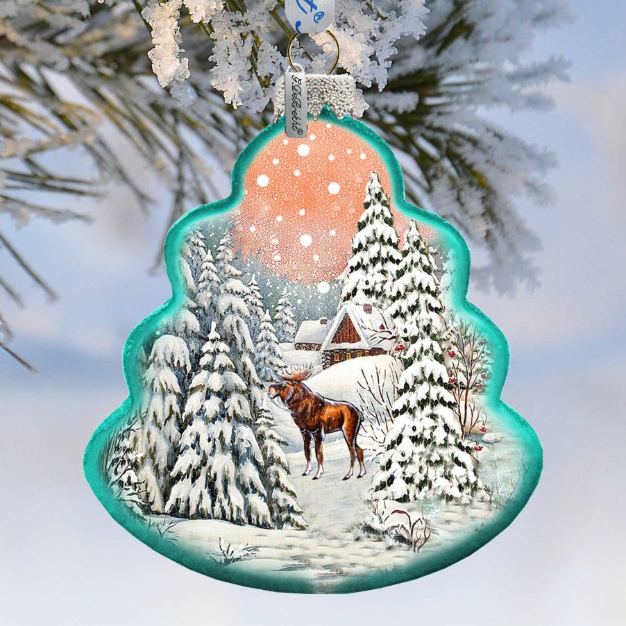 A decorative ornament hanging from a tree branch, depicting a snowy winter scene with a moose in the foreground and a snowy forest in the background.