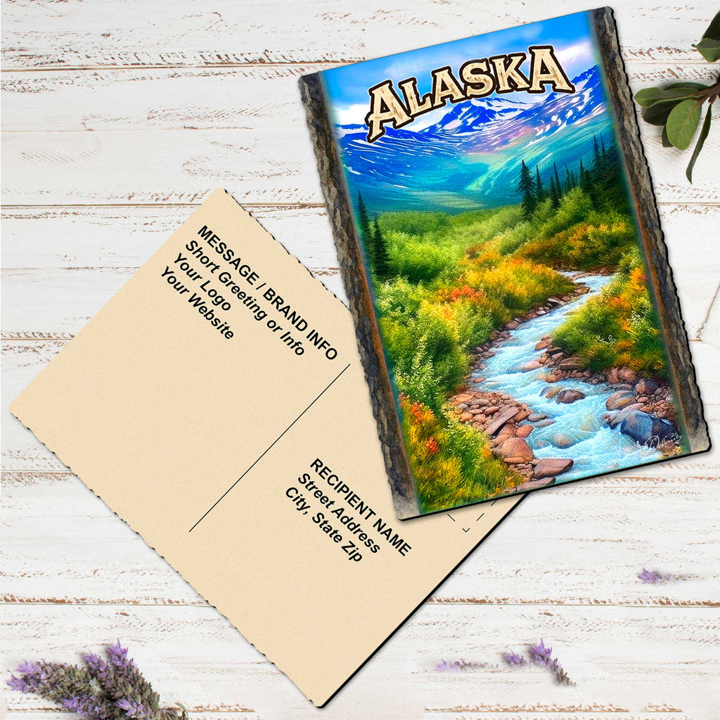A postcard featuring a scenic landscape of a river flowing through a forest, with mountains in the background. The postcard is placed on a wooden surface, and there are some purple flowers nearby.