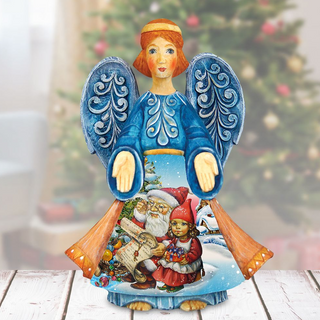 A figurine of an angel with wings, wearing a blue dress and holding a Christmas ornament, is standing on a wooden surface in front of a blurred Christmas tree.