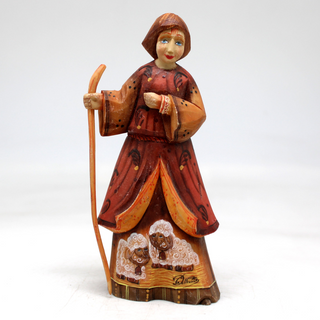 A wooden figurine depicting a woman in a red dress and holding a walking stick, with two sheep on her dress.