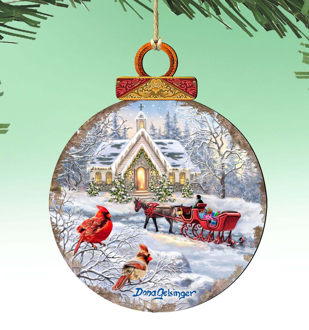 A round ornament depicting a snowy winter scene with a church, a horse-drawn sleigh, and two red cardinals perched on a tree branch.