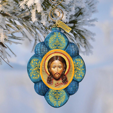 A blue and gold ornament featuring a depiction of Jesus Christ is hanging from a branch with snow on it.