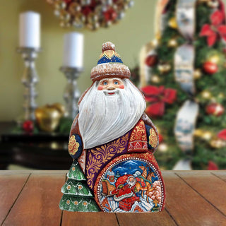 A wooden Santa Claus figurine with intricate carvings and decorations sits on a wooden surface, with a blurred Christmas tree and other holiday decorations in the background.