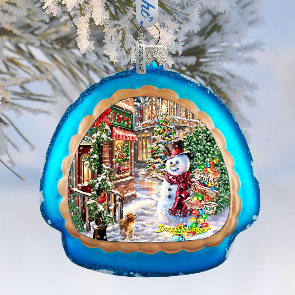 A blue ornament with a snowman and a dog in a snowy winter scene, hanging on a tree branch.