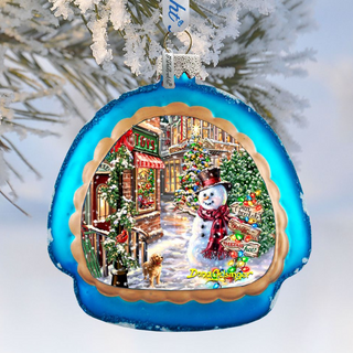 A blue ornament with a snowman and a dog in a snowy winter scene, hanging on a tree branch.