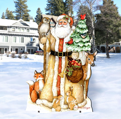 A wooden cutout of Santa Claus holding a Christmas tree, a bag of presents, and various animals such as a fox, a rabbit, and a deer, set against a snowy background with a house and trees.