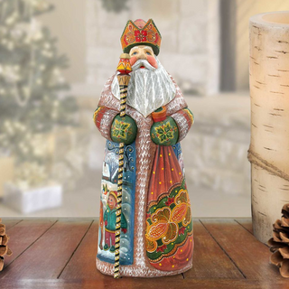 A colorful, hand-painted figurine of a saint or religious figure, wearing a long robe and holding a staff, stands on a wooden surface.
