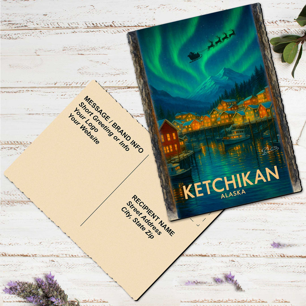 A postcard featuring a scenic image of a town in Alaska with a snowy mountain in the background and a sleigh flying over the town. The postcard is placed on a white wooden surface, with some purple flowers nearby.