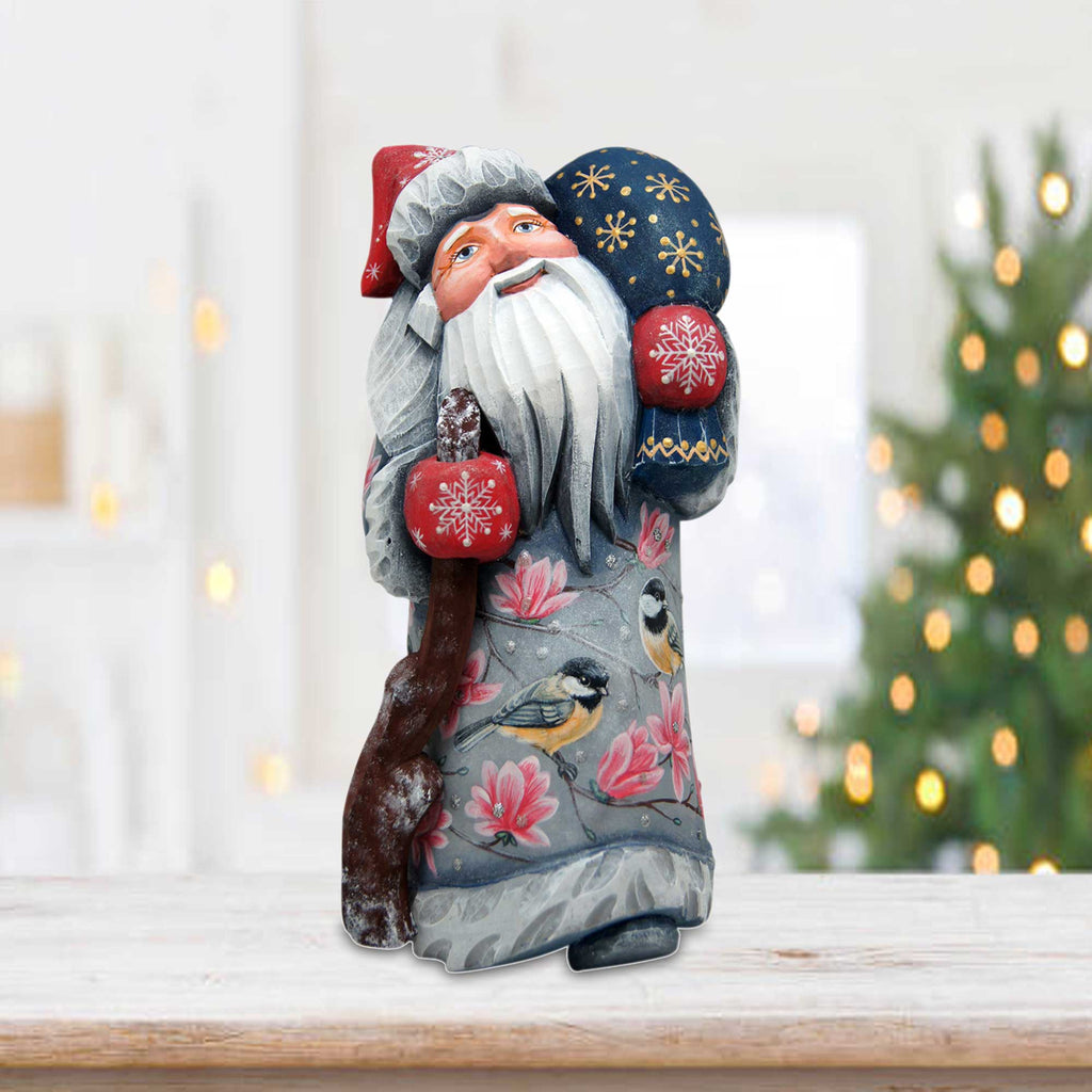 A wooden Santa Claus figurine with a blue hat and red mittens is sitting on a wooden surface, with a blurred Christmas tree in the background.