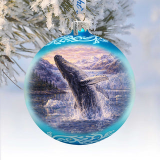 A blue ornament with a painting of a whale jumping out of the water, surrounded by snow-covered pine branches.