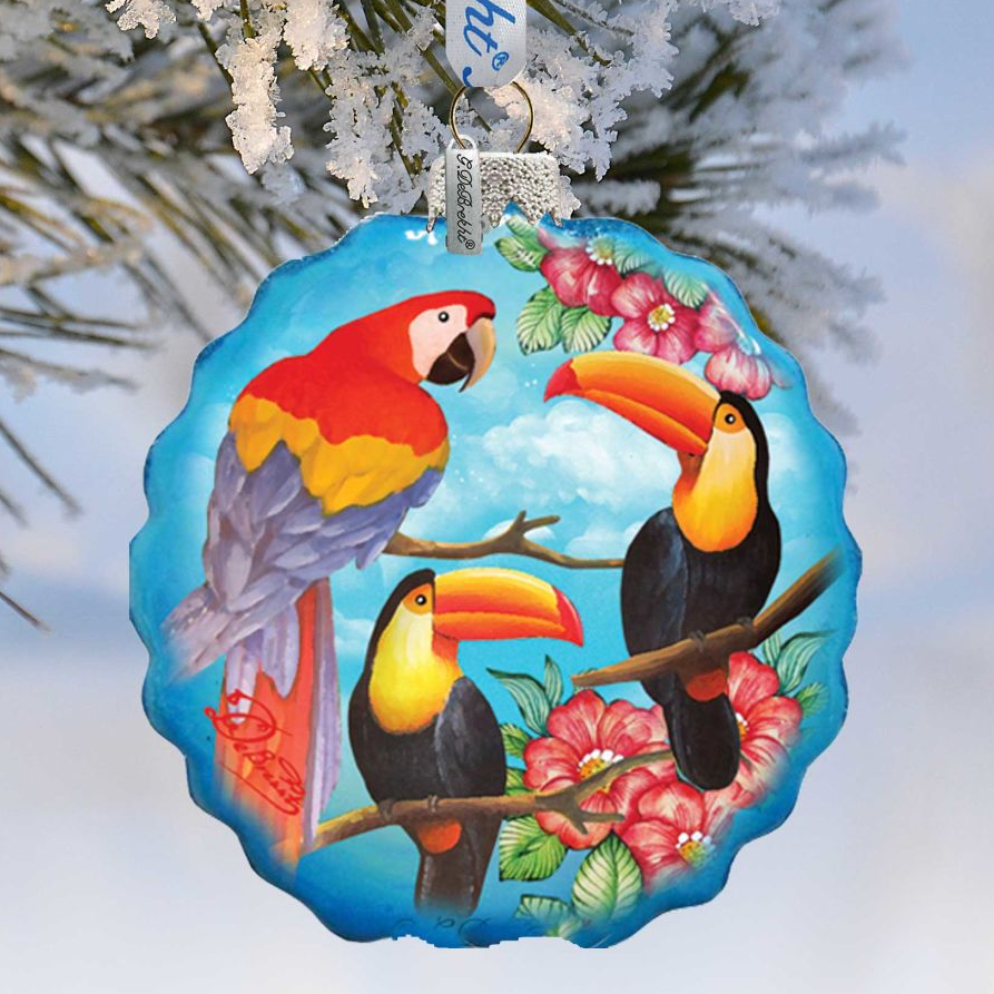 A blue ornament with a tropical bird design, featuring a parrot, a toucan, and a macaw, hanging on a tree branch.