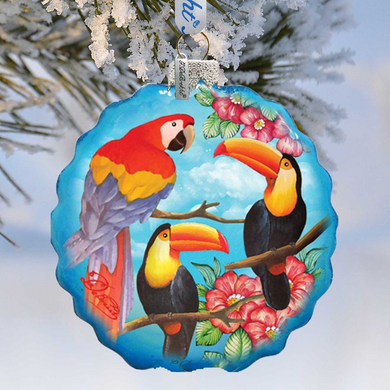 A blue ornament with a tropical bird design, featuring a parrot, a toucan, and a macaw, hanging on a tree branch.