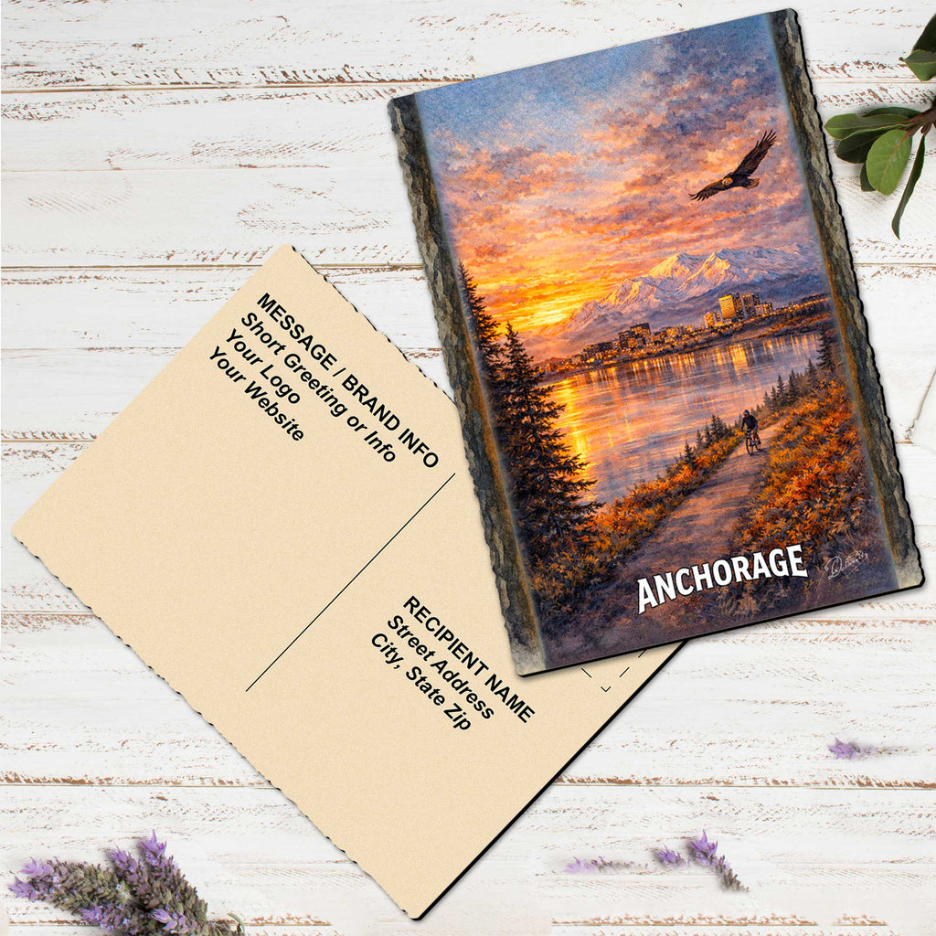 a postcard with a painting of a scenic landscape, including mountains, a river, and a sunset. The postcard is placed on a white wooden surface, with some purple flowers nearby.