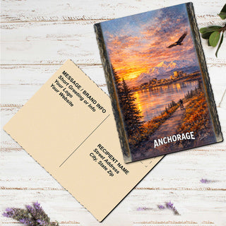 a postcard with a painting of a scenic landscape, including mountains, a river, and a sunset. The postcard is placed on a white wooden surface, with some purple flowers nearby.
