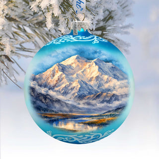 A blue Christmas ornament with a painting of a snowy mountain landscape, featuring a lake and snow-covered peaks, hanging from a tree branch.