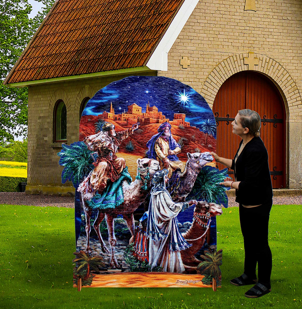 A person is standing next to a large, colorful, and detailed cardboard nativity scene depicting three figures on camels, with a castle and starry sky in the background.