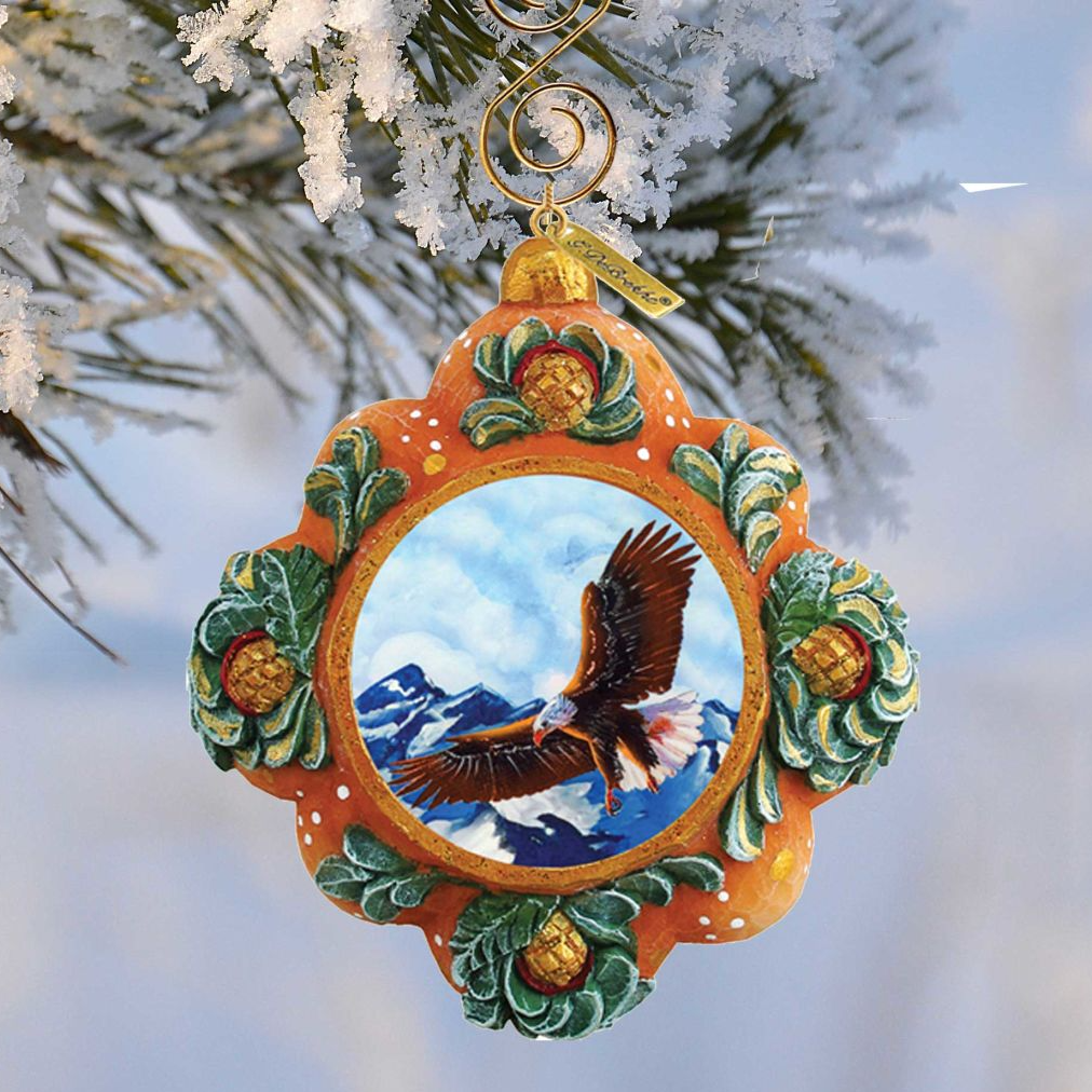A decorative ornament hanging from a tree branch, featuring a painted eagle in flight against a snowy mountain backdrop.
