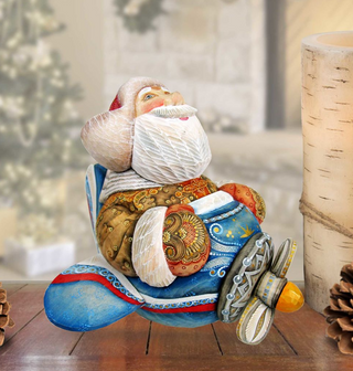 A colorful, handcrafted wooden Santa Claus figurine sitting on a blue and red airplane-shaped base, placed on a wooden surface.