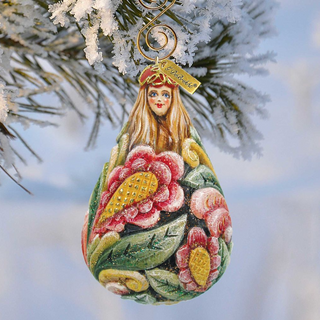 A colorful, hand-painted Christmas ornament with a woman's face and flowers hangs from a tree branch against a snowy background.