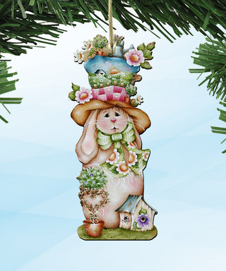 A whimsical ornament depicting a rabbit wearing a hat and holding a basket of flowers, with a small house and tree in the background.