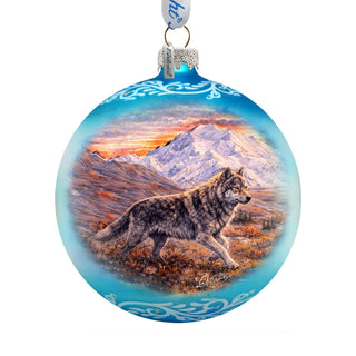 A blue glass ornament with a painting of a wolf walking through a mountainous landscape at sunset.