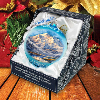 A blue Christmas ornament with a mountain landscape is displayed in a black and white box, surrounded by holiday decorations.