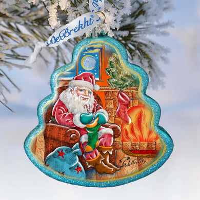 A Christmas ornament depicting Santa Claus sitting in a chair by a fireplace, with a fire burning and a bag of presents next to him.
