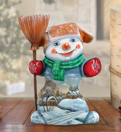A snowman figurine with a painted winter landscape on its body, holding a broom and wearing a green scarf.
