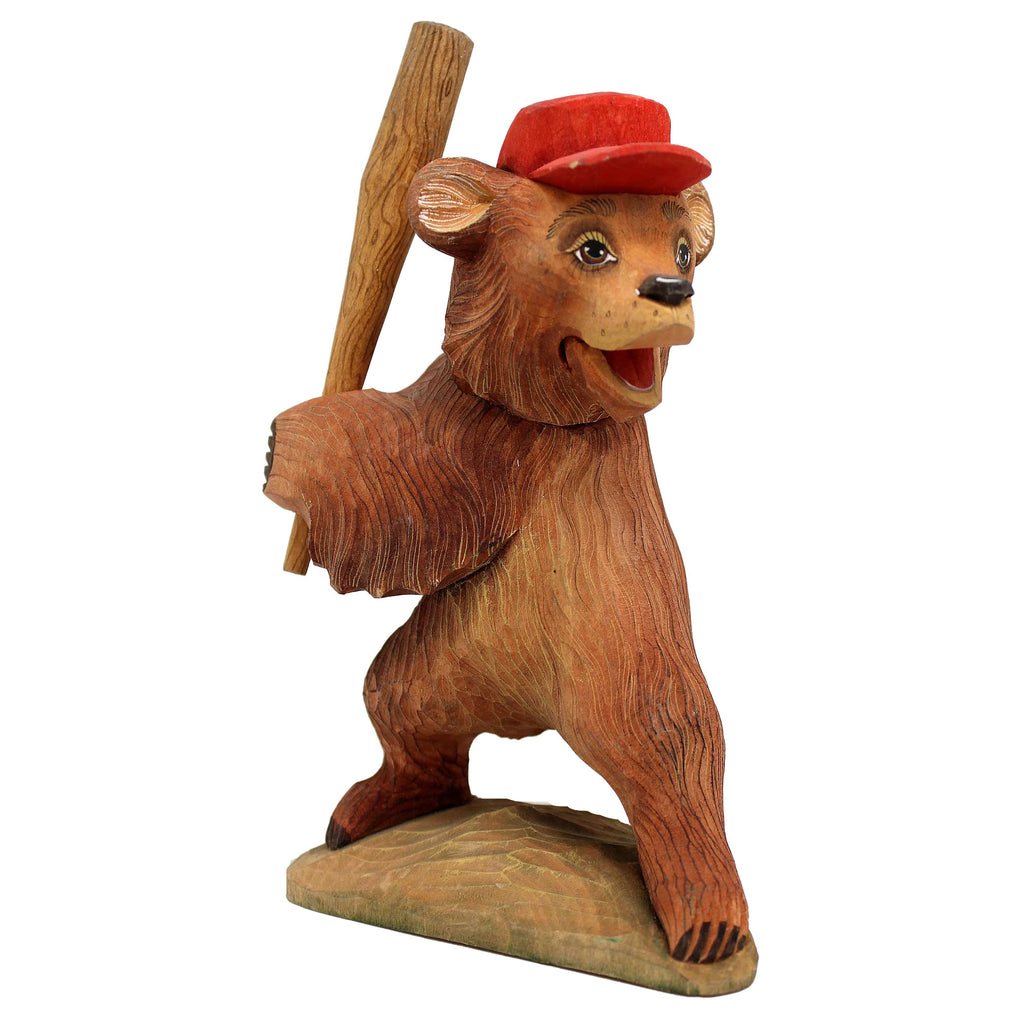 A wooden sculpture of a bear wearing a red baseball cap and holding a baseball bat.