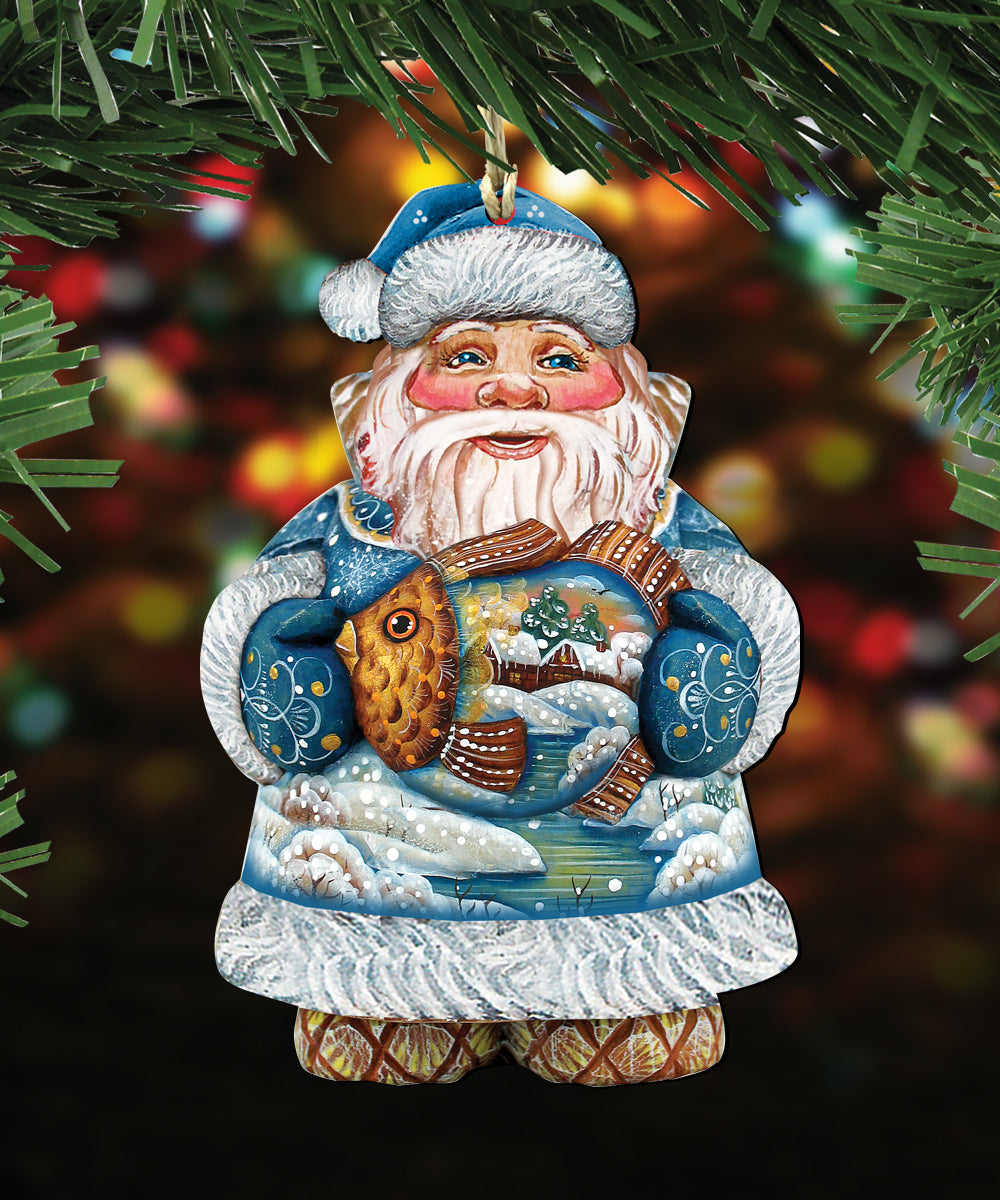 A Christmas ornament in the shape of Santa Claus, with a blue hat, white beard, and holding a fish. The ornament is hanging from a tree branch with green pine needles.