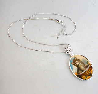 A silver necklace with a pendant featuring a small kitten sitting in a basket.