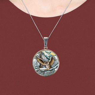 A silver pendant necklace with a circular design featuring an owl in flight against a snowy background.