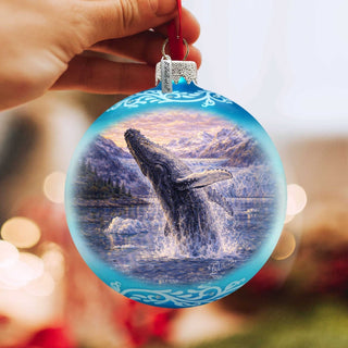 A hand holds a blue ornament with a painting of a whale leaping out of the water, set against a backdrop of mountains and a sunset sky.