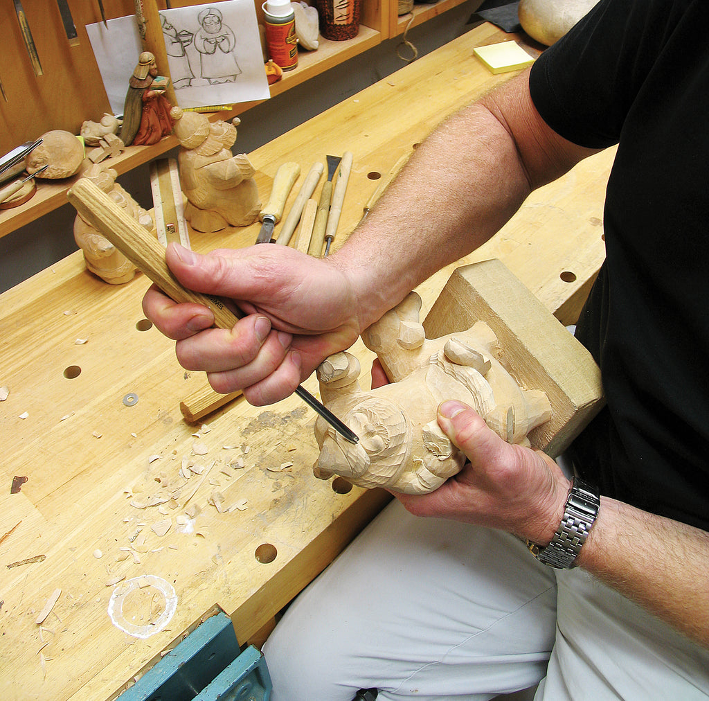 A man is carving a wooden sculpture in a workshop, using various tools to shape the wood.