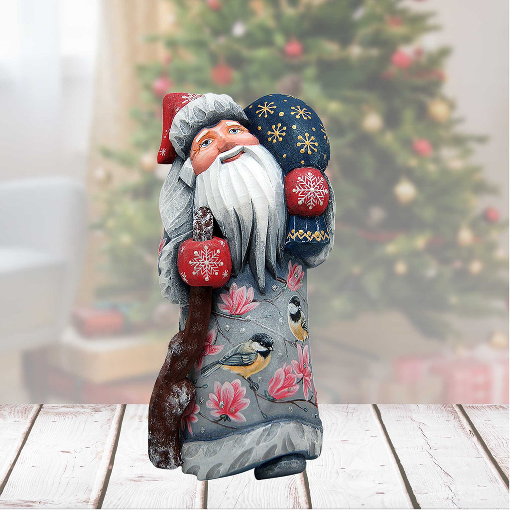 A wooden Santa Claus figurine holding a red snowflake ornament and a blue snowflake ornament, standing in front of a Christmas tree.