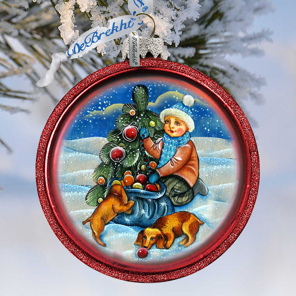 A red ornament with a Christmas tree and a boy in a hat, along with a dog, is hanging on a tree branch.