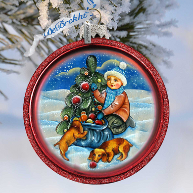 A red ornament with a Christmas tree and a boy in a hat, along with a dog, is hanging on a tree branch.