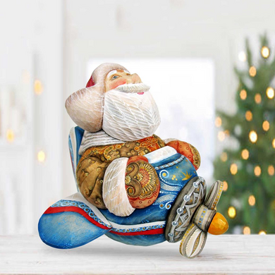 A colorful, hand-painted Santa Claus figurine sitting on a blue and red airplane-shaped base, with a blurred Christmas tree in the background.